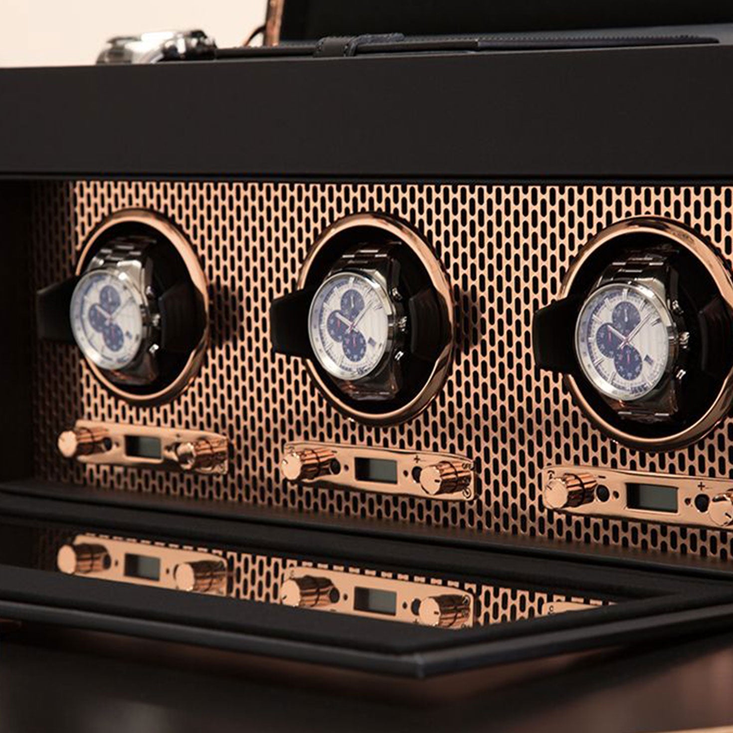 The WOLF 1834 Axis Watch Winder by WOLF stylishly displays three luxury watches with a sophisticated black and copper finish. Each watch is safely nestled in its own compartment, set against a grid pattern backdrop with control knobs beneath, all housed within a sleek, modern cabinet perfect for discerning watch enthusiasts.