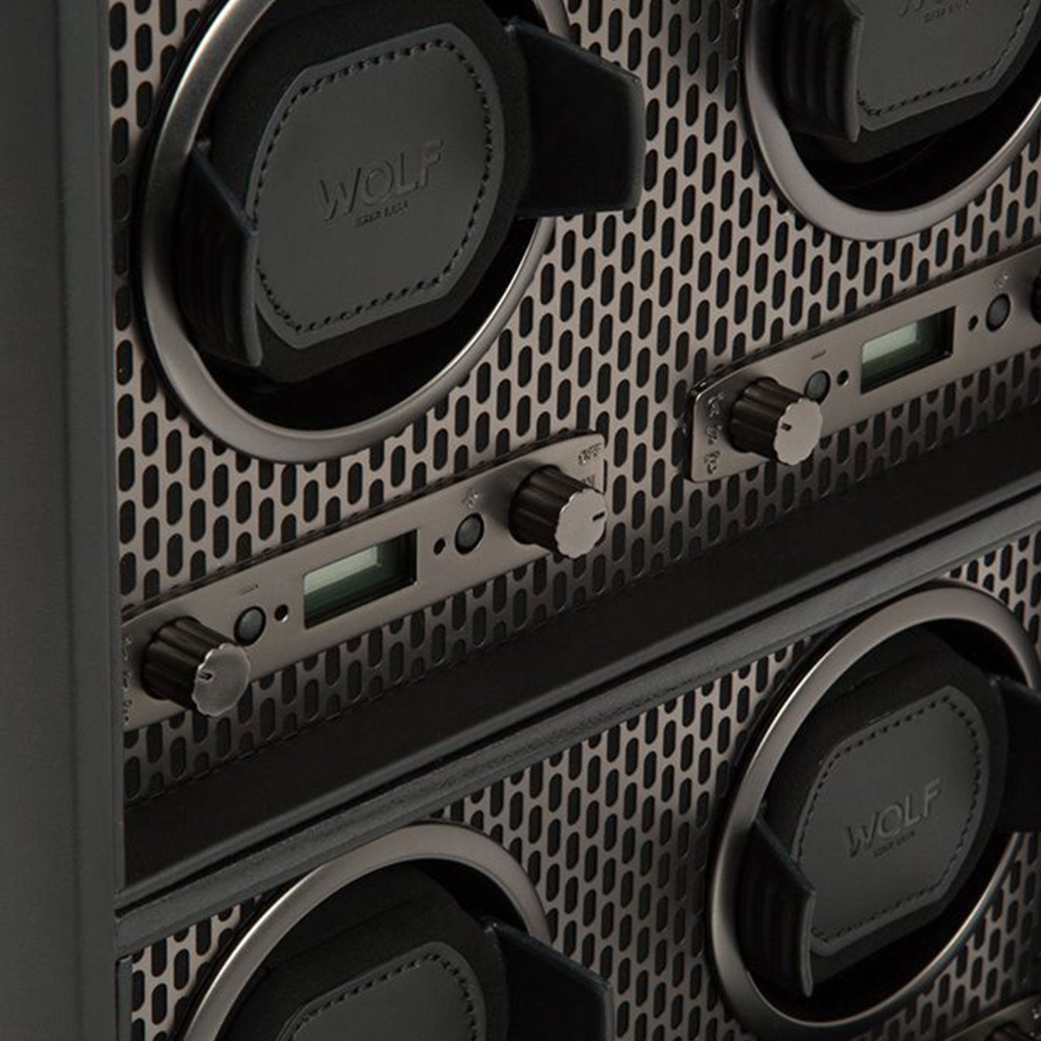 A close-up of the WOLF 1834 Axis 4 Piece Winder reveals a luxury timepiece storage solution with four compartments. Each section is equipped with a black leather pad, digital display, and dials for rotation options. The metal panel showcases a perforated design, and the brand name "WOLF" is prominently visible on the pads.