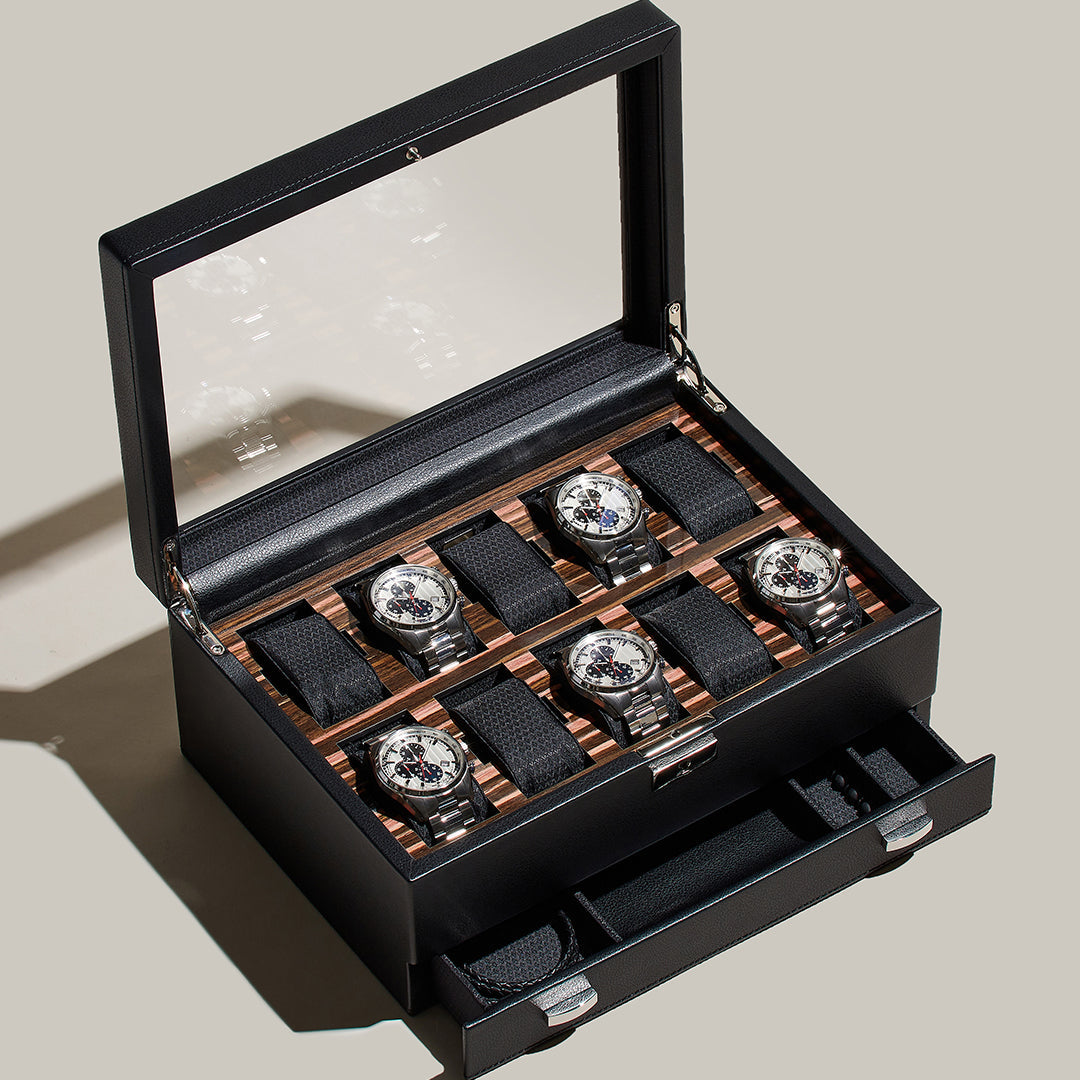The WOLF 1834 Roadster 10 Piece Watch Box With Drawer features a leather exterior with a glass lid, securely holding six wristwatches on black cushions. Its wood-like finish interior provides optimal watch storage, while a lower drawer accommodates six extra watch straps. The box is displayed in a well-lit setting and includes lock and key security for enhanced protection.