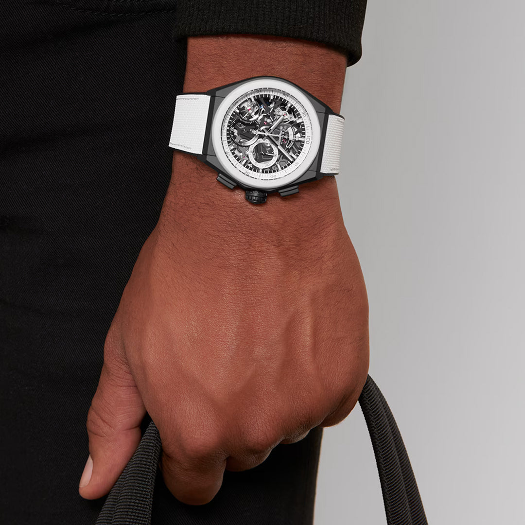 A close-up reveals a hand gripping a black strap, wearing a Zenith Defy El Primero 21 Watch with white strap and an intricate black and silver dial in a sleek ceramic 44mm case, set against a plain gray background.