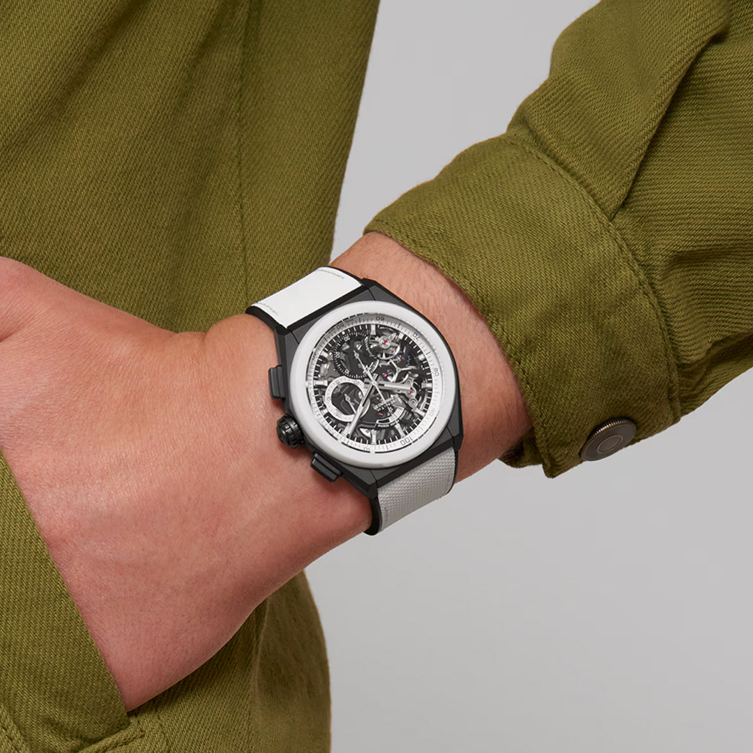 A person in a green jacket showcases the ZENITH Defy El Primero 21 El Primero 9004, featuring a ceramic case, white and black strap. The transparent dial reveals intricate gears of the precise El Primero 21 chronograph inside.