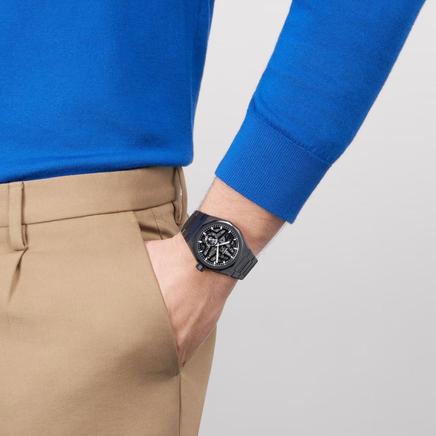 A person in a bright blue long-sleeve shirt and tan pants has their hand in their pocket. They are wearing the ZENITH Defy Skyline Skeleton El Primero 3620 SK Automatic 41mm Watch, featuring a sleek black ceramic case with the intricate movement visible on its dial.