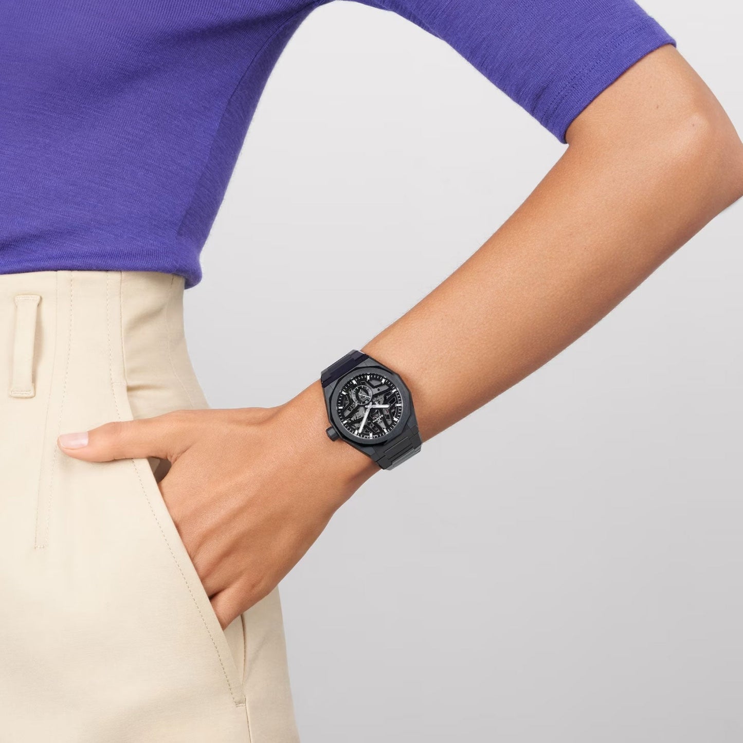 A person dressed in a purple shirt and beige pants displays the ZENITH Defy Skyline Skeleton El Primero 3620 SK Automatic 41mm Watch. This watch, designed by Zenith, boasts a round and stylish aesthetic with an intricate dial and dark band, all highlighted against a neutral background.