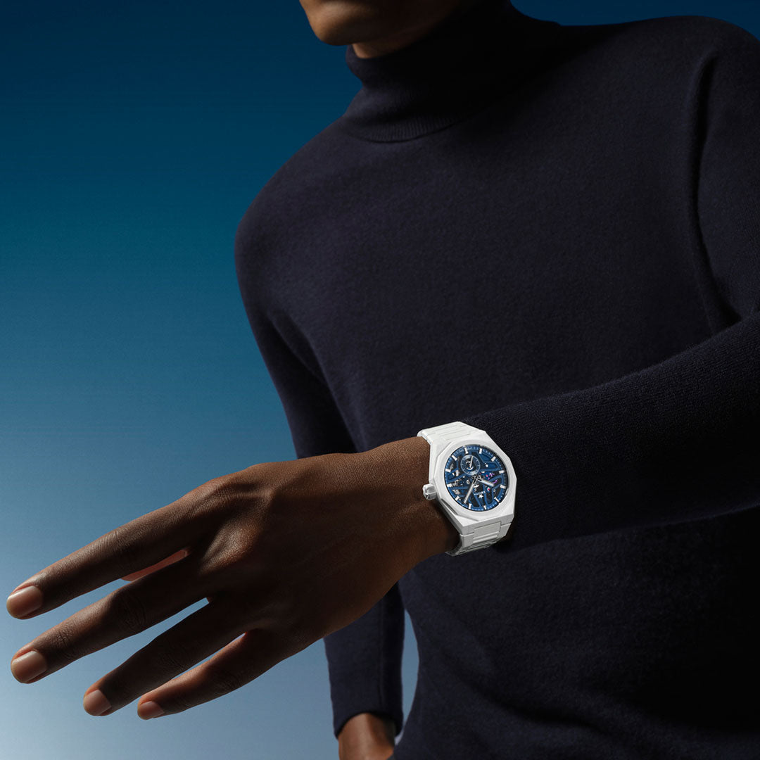 A person wearing a dark turtleneck sweater showcases the ZENITH Defy Skyline Skeleton El Primero 3620 SK Automatic 41mm Watch, featuring a blue face and silver strap. The background is a gradient of blue hues that perfectly complements the watch's elegance, with the focus remaining on the timepiece and the person's outstretched arm.