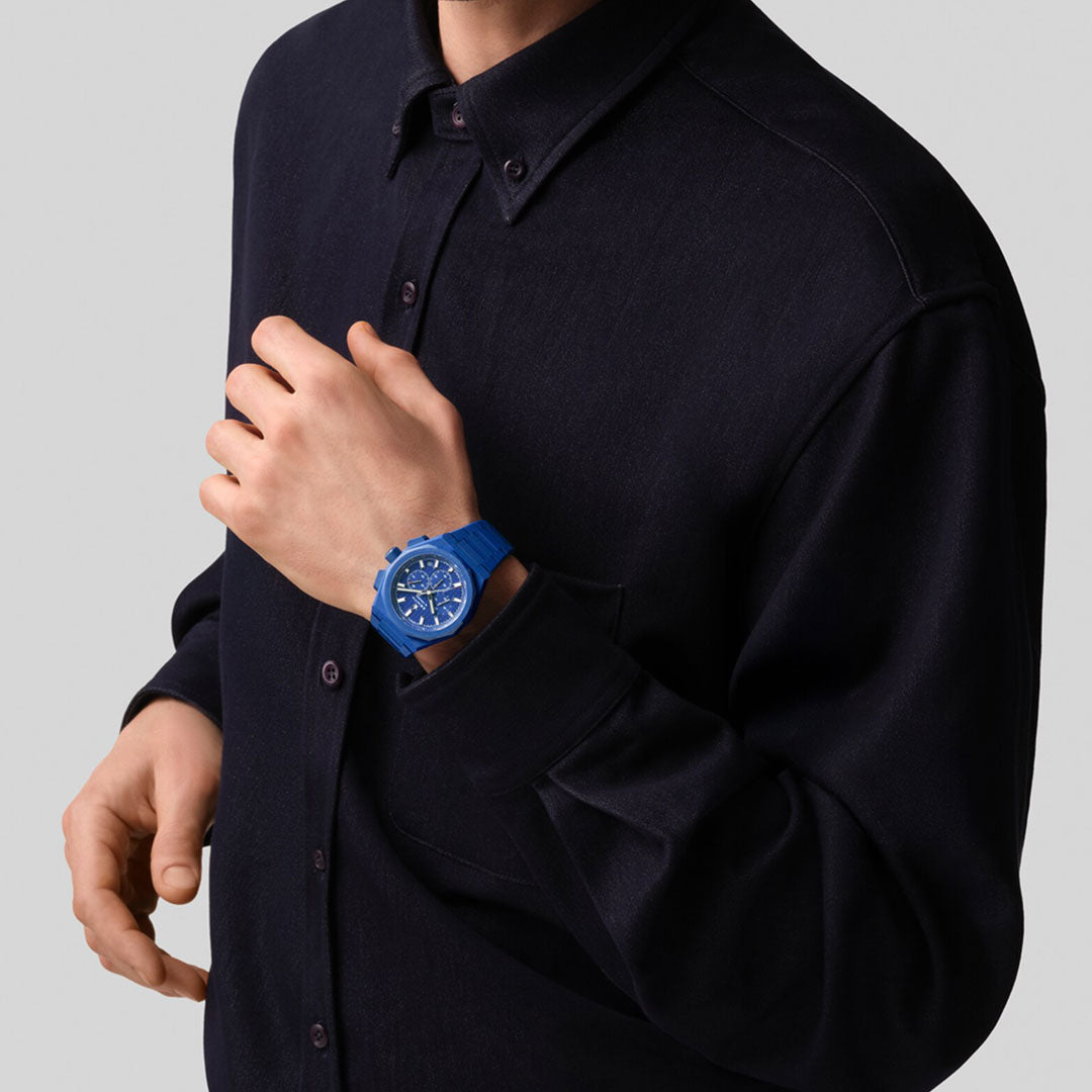 A person in a dark button-up shirt wears the Zenith Defy Skyline Chronograph 160th Anniversary Limited Edition El Primero 3600 42mm watch, its bold blue face clearly visible as their left hand touches their chest against a light gray background.