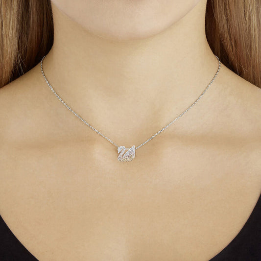 A woman is wearing the Swarovski Iconic Swan Necklace Pendant, which features a swan-shaped design adorned with dazzling Swarovski Crystals. The Rhodium-plated piece from Swarovski rests gracefully at the base of her neck on a neutral background.