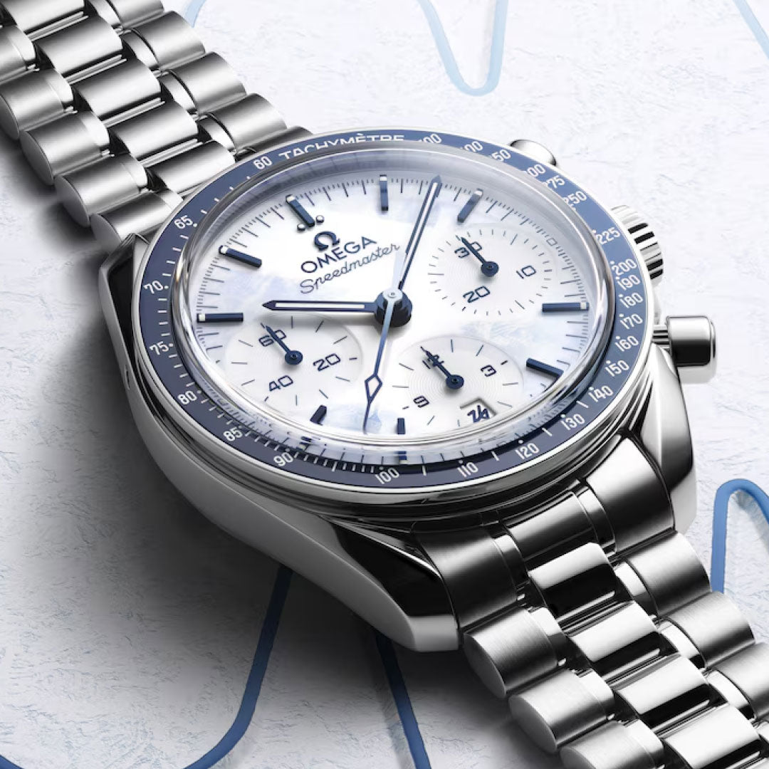 A close-up of the OMEGA Speedmaster 38 Co-Axial Chronometer Chronograph Milano Cortina 2026 Edition, featuring a stainless steel bracelet, white dial with blue accents and three subdials, set against a light background with subtle blue lines.