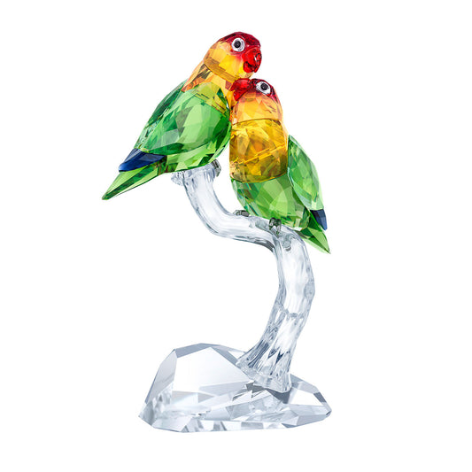 The Swarovski Lovebirds Home Décor is a captivating crystal sculpture that showcases two vividly colored lovebirds perched on a clear, polished branch. These birds boast green bodies, yellow necks, and red heads crafted from faceted glass, offering a vibrant and reflective look signature to Swarovski's design.