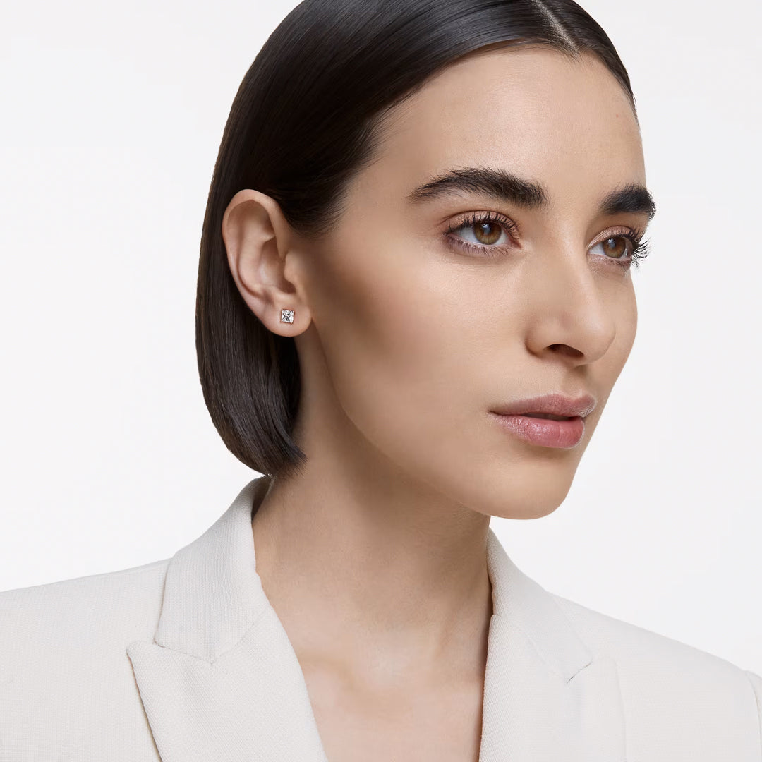 A woman with short, dark hair is wearing the Swarovski Stilla Attract stud earrings in rose gold and a white blazer. She gazes to the side, exuding a unisex aesthetic against a plain white background.