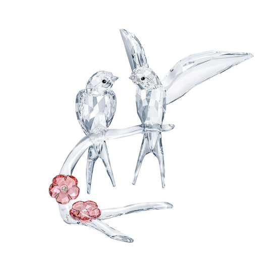 The Swarovski Swallows Home Décor piece features two exquisitely detailed birds resting on a branch with pink crystal flowers. Masterfully crafted to capture and reflect light, it is a perfect romantic gift for any nature-inspired collection.