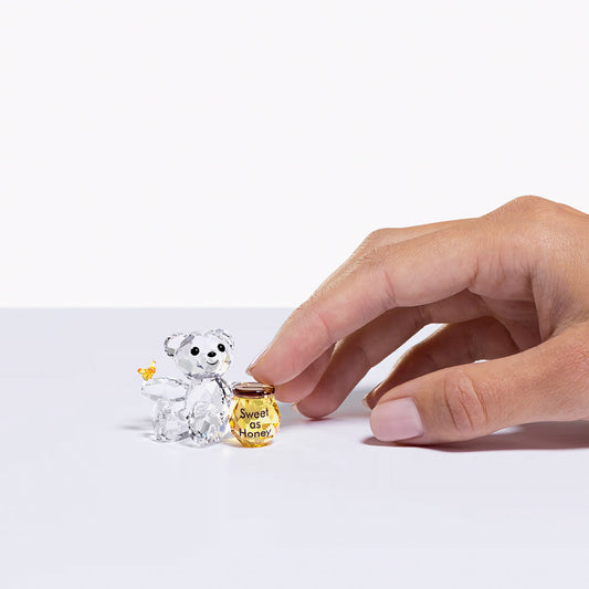 A hand carefully sets down a miniature amber-colored "Sweet as Honey" jar next to the Swarovski Kris Bear Sweet as Honey Home Décor crystal figurine, featuring a small bee on its ear. The scene is elegantly displayed against a plain white background, showcasing the exquisite craftsmanship of the clear crystal piece by Swarovski.