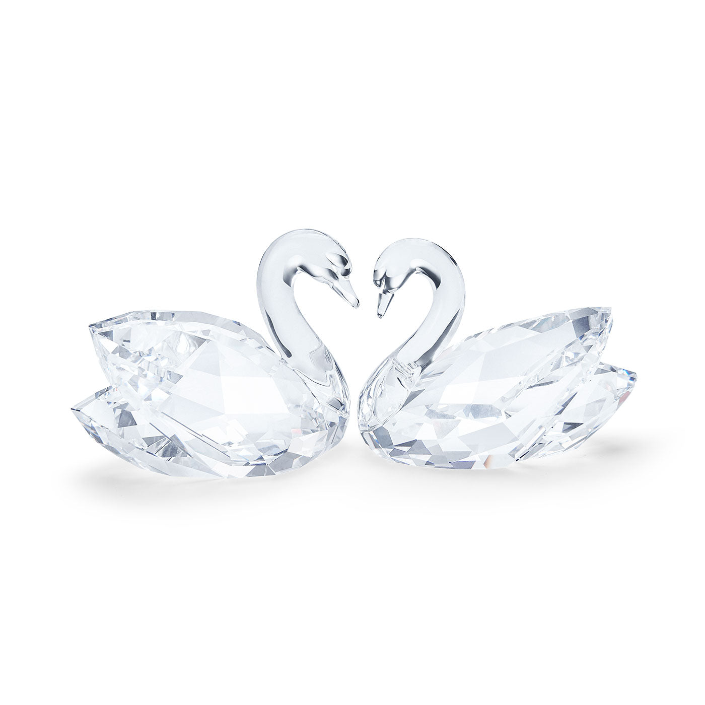 The Swarovski Swan Couple Home Décor features two exquisite crystal swans poised towards one another, their necks gracefully arching to create a romantic heart shape. Crafted by Swarovski, these pieces have faceted surfaces that beautifully capture the light, highlighting their transparent allure against a simple white backdrop.