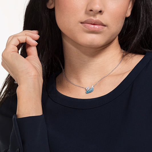 A woman wearing a navy blue top and a rhodium-plated necklace elegantly adorned with blue crystals. Her hand rests gently near her neck, subtly showcasing the Swarovski Iconic Swan Necklace Pendant by Swarovski, as she gazes slightly off-camera against a softly lit, plain background.