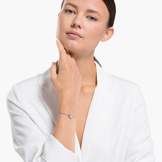 A person dressed in a white blazer poses against a simple backdrop, their hair sleekly pulled back. With one hand elegantly touching their chin, they display the rhodium-plated Swarovski Attract Soul Bracelet, featuring a purple gemstone, on their wrist.