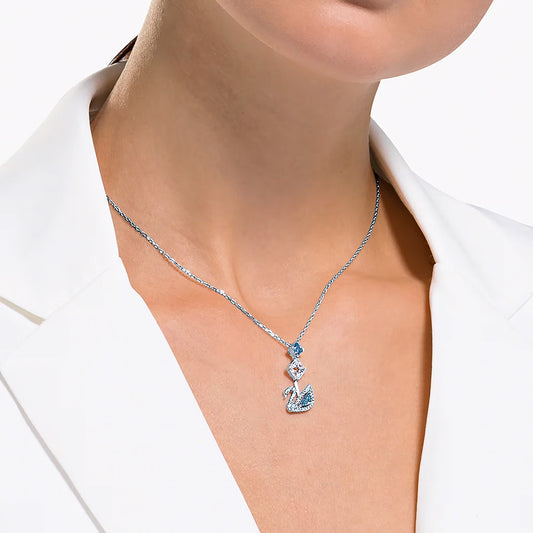 Dressed in a white blazer, an individual models the Swarovski Swan Necklace Pendant, which boasts a silver chain and a striking swan pendant embellished with blue and clear gemstones. This elegant necklace, featuring Swarovski's iconic swan logo, is ideal for special occasions.