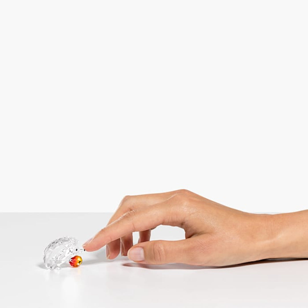 A hand gently touches the Swarovski Hedgehog with Apple Home Décor, featuring a small, translucent design and a red and yellow snout, as it rests on a white surface against a plain background.