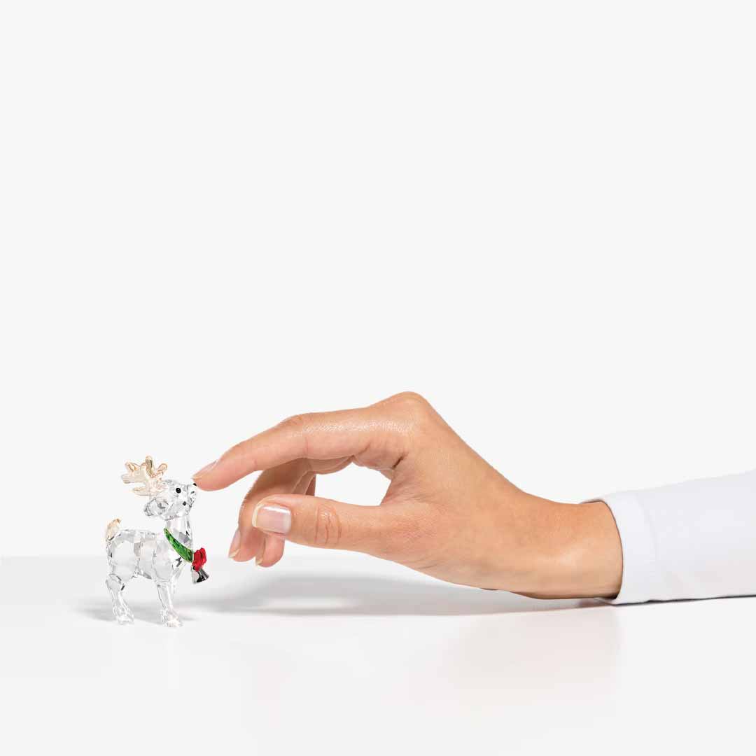 A hand delicately touches a small, intricately designed Swarovski Santa's Reindeer Home Décor piece featuring crystal antlers and a green scarf. The festive decoration holds a red rose, positioned on a smooth, white surface against a plain background.