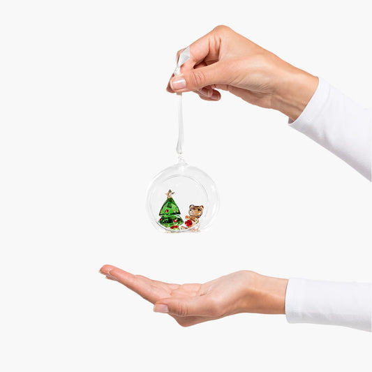 A person holds the Swarovski Joyful Ball Christmas Scene Ornament Home Décor suspended from a ribbon. Encased within the transparent ornament are tiny decorative elements: a festive tree and a small teddy bear. The display is set against a plain white background, perfectly capturing the essence of holiday charm.