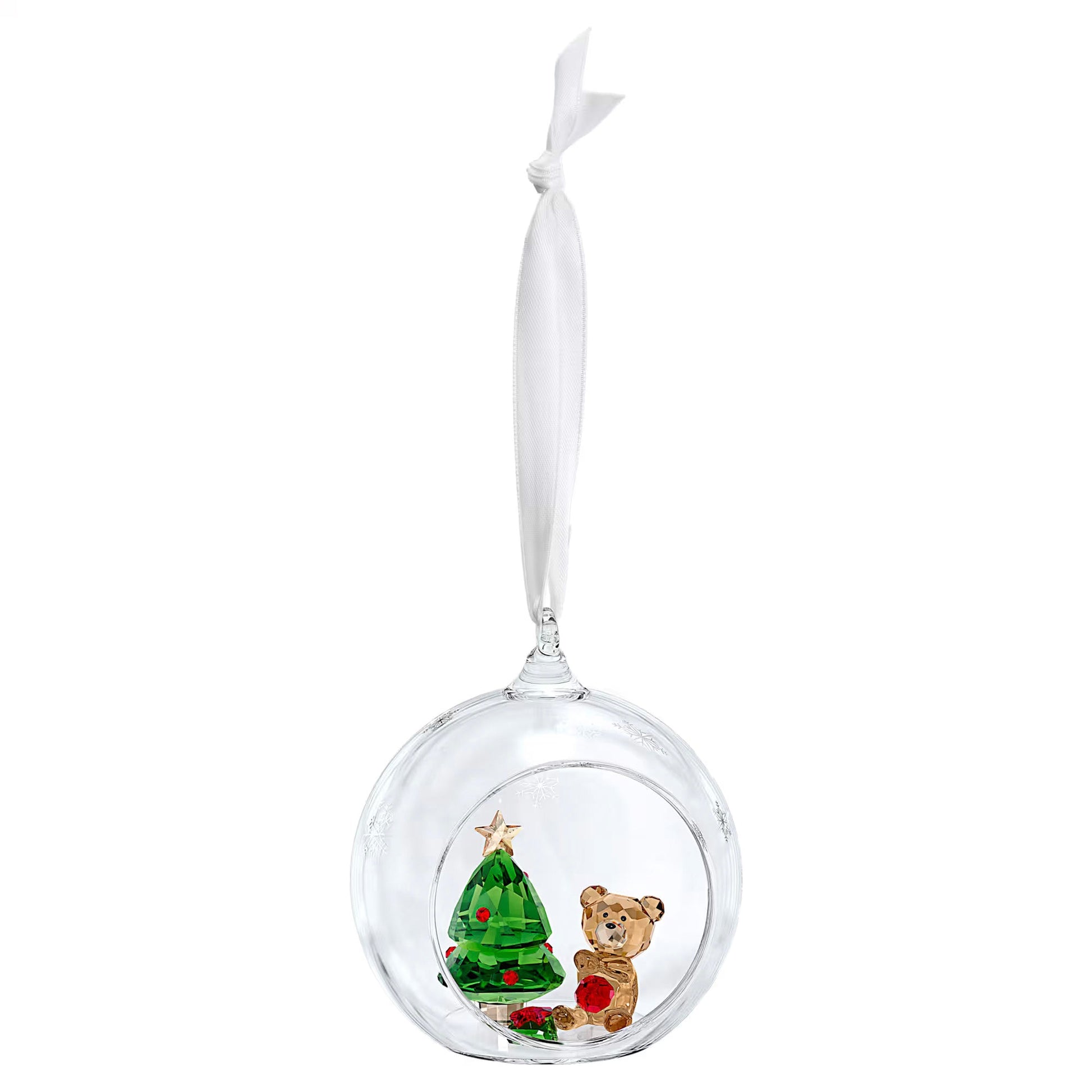 The Swarovski Joyful Ball Christmas Scene Ornament from the renowned brand, Swarovski, features a transparent ornament encasing a small green crystal Christmas tree topped with a gold star. Accompanying this scene is an adorable brown crystal teddy bear holding a gift, all gracefully hanging from a white ribbon to enhance the elegance of your holiday décor.