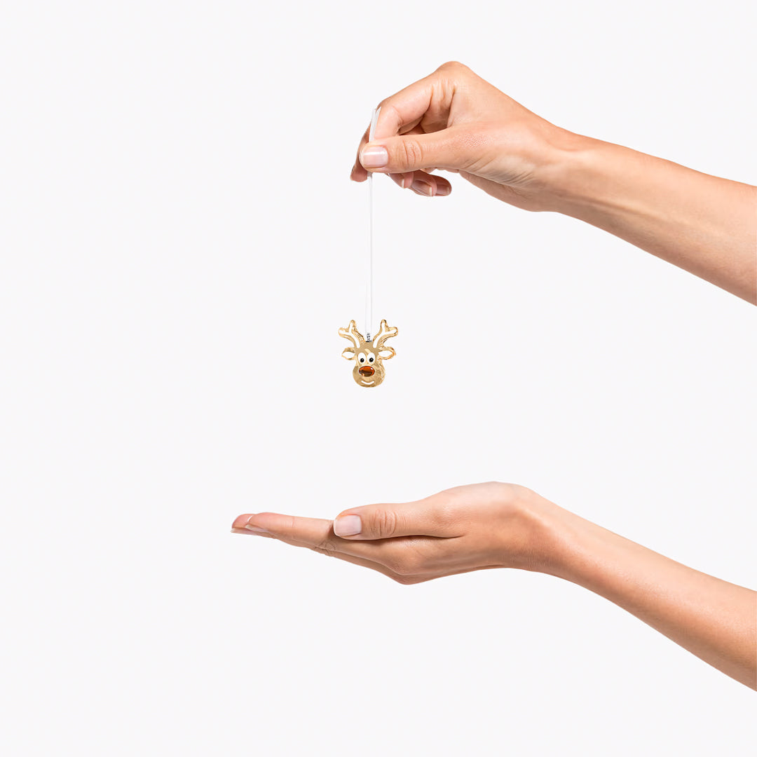 A person's hand holds the Swarovski Gingerbread Reindeer Ornament Home Décor, a small, gold piece accented with sparkling crystal, and suspended by a string above their open palm. The background is plain white.