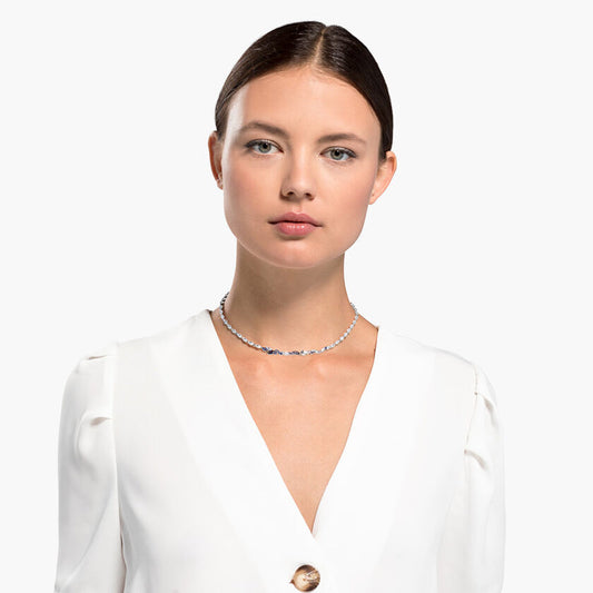 A woman with a neutral expression is wearing a white blazer and the Swarovski Louison Necklace, crafted by Swarovski and embellished with small blue stones. The background is plain white.