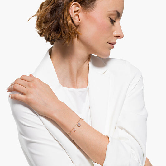 A woman with short brown hair, dressed in a white blazer and top, poses with her arms crossed while gazing downward, highlighting a delicate Swarovski Magic Bracelet in rose gold-tone on her wrist. The background is plain white.
