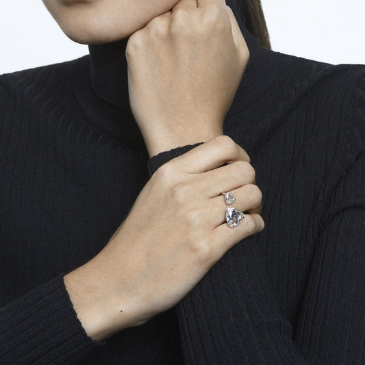 A person wearing a black turtleneck sweater displays their hand adorned with two large, sparkling Swarovski Millenia Cocktail Rings, which feature prominent stones. Their other hand rests thoughtfully against their chin, with the plain white background emphasizing the elegance of the trilliant cut crystals from Swarovski.
