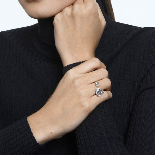 Dressed in a black turtleneck sweater, an individual is showcasing the dazzling Swarovski Millenia Open Ring on their right hand. This elegant ring features a trilliant-cut white crystal set in rhodium-plated metal. With their left hand near their face, the shimmering beauty of the ring is accentuated.