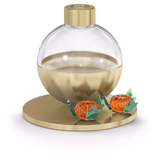 A Swarovski Garden Tales Pumpkin Scent Diffuser Container, with its elegant round glass design and gold base, sits gracefully on a matching circular gold platform, exuding the luxury associated with Swarovski. Beside it are two orange, translucent wrapped candies that enhance the opulent display with a sweet touch.