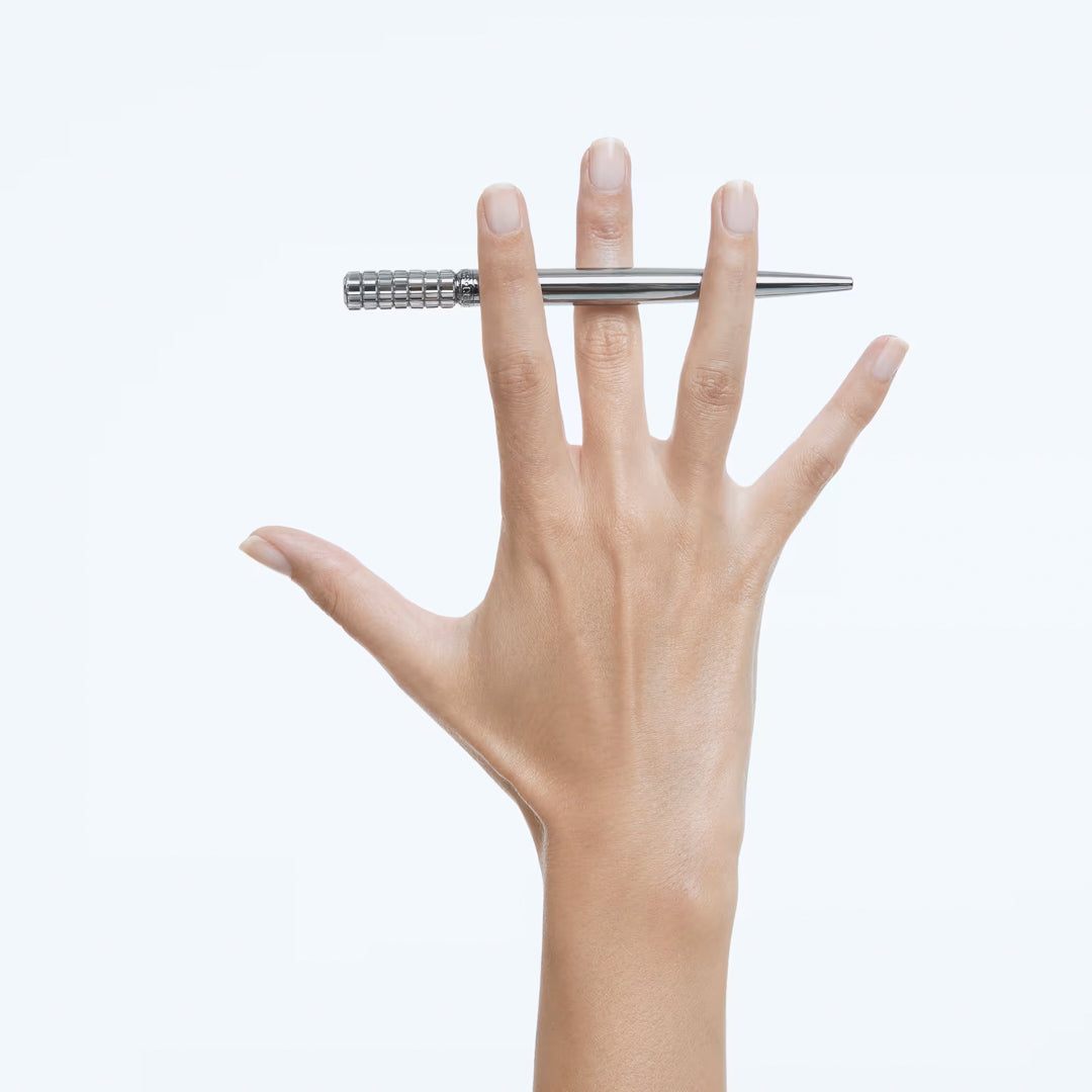 A hand with fingers spread apart holds a sleek Swarovski Ballpoint Pen, its crystal design glistening as it's balanced between the middle and ring fingers against a plain white background.