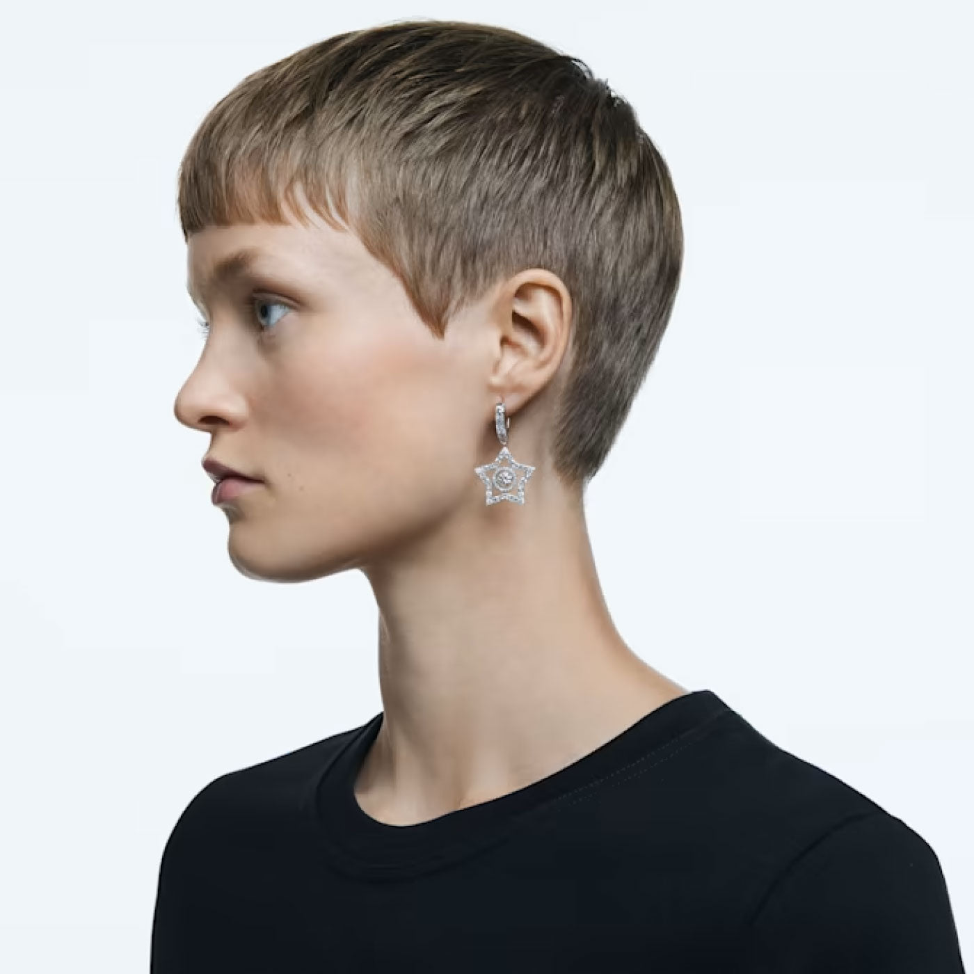 A person with short hair is shown in profile, wearing a Swarovski Mixed Cuts Star Stella Drop Earring that features a round gemstone. They are dressed in a black top set against a light grey background.