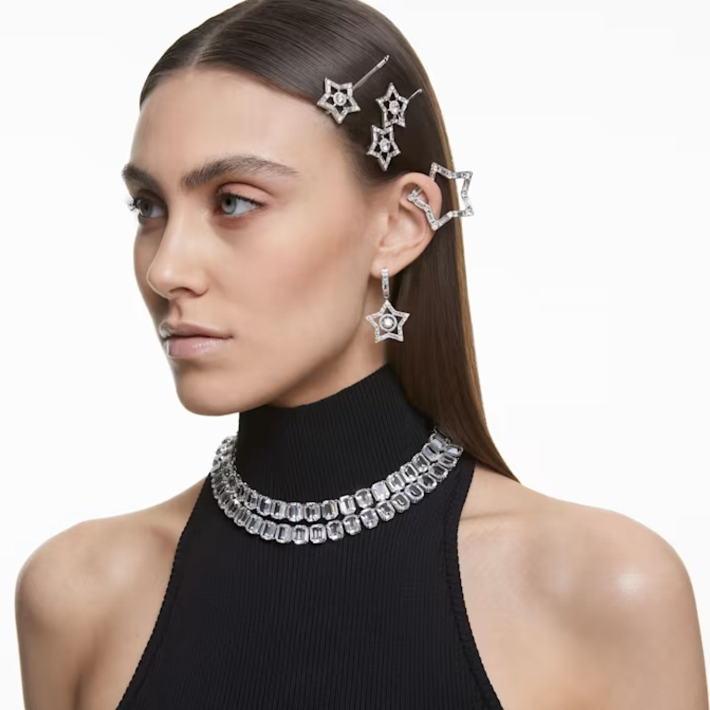 A woman with straight brown hair wears a black high-neck sleeveless top. Embracing Stella family fashion, she accessorizes with Swarovski's Mixed Cuts Star Stella Drop Earrings, star-shaped hairpins, and a gemstone-adorned choker, all set against a plain white background.