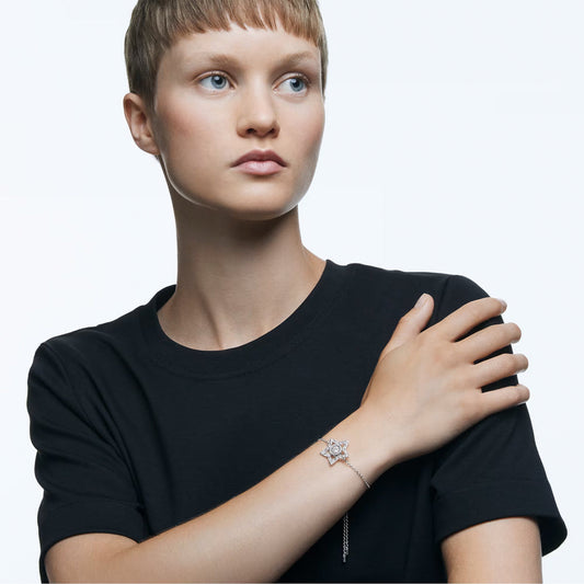 A person with short hair, dressed in a black t-shirt, poses against a plain background while showcasing the elegant Swarovski Stella Bracelet by Swarovski. The bracelet features a star-shaped charm and a dancing stone on their wrist. Their right hand rests gracefully on their left shoulder, highlighting the accessory's sophistication.
