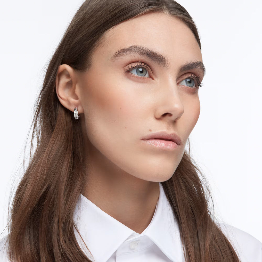 A person with long brown hair and light eyes gazes upward, wearing a white collared shirt and the shimmering Swarovski Dextera Hoop Earrings. The background is a plain white.