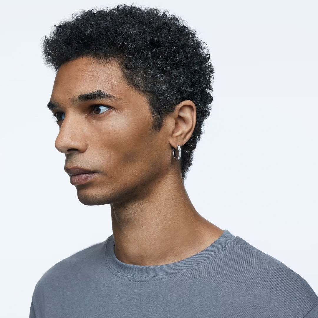 A person with short curly hair, dressed in a gray t-shirt and adorned with the Swarovski Dextera Hoop Earrings, gazes to the side against a simple backdrop.
