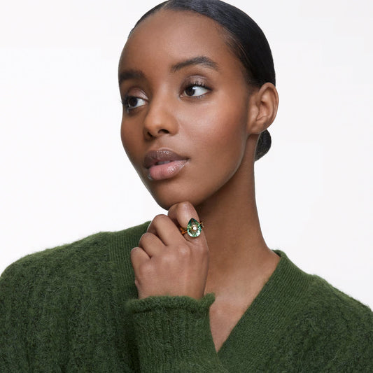 A woman wearing a green sweater gazes off to the side, resting her chin on her hand that showcases the Swarovski Numina Ring. This exquisite piece from Swarovski features an intricate design centered around a striking green crystal. The plain white background accentuates not only her serene expression but also the ring's elegance perfectly.