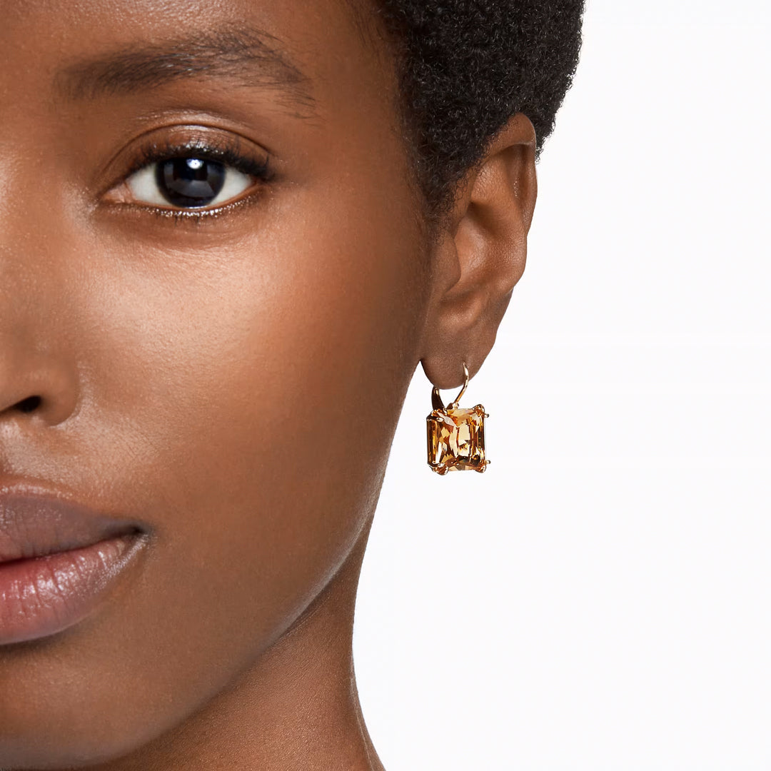 Close-up of a person wearing Swarovski Millenia Drop Earrings, which feature a gold-tone plated abstract design adorned with sparkling square-cut crystals. The earrings are prominently displayed against a neutral background, highlighting their intricate shape and shiny finish. The person has a smooth complexion and short black hair.