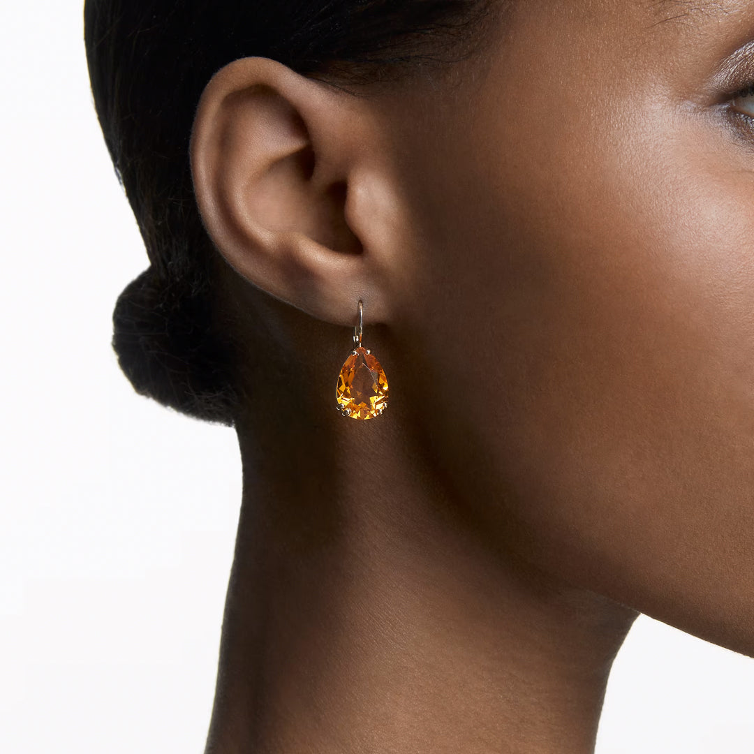 An individual is seen wearing Swarovski Millenia Drop Earrings, showcasing a teardrop-shaped orange gemstone that stands out beautifully against their skin. The jewelry's sophistication is enhanced by the gold-tone plated metal and yellow crystals, with hair neatly pulled back to highlight the earrings' radiant charm.