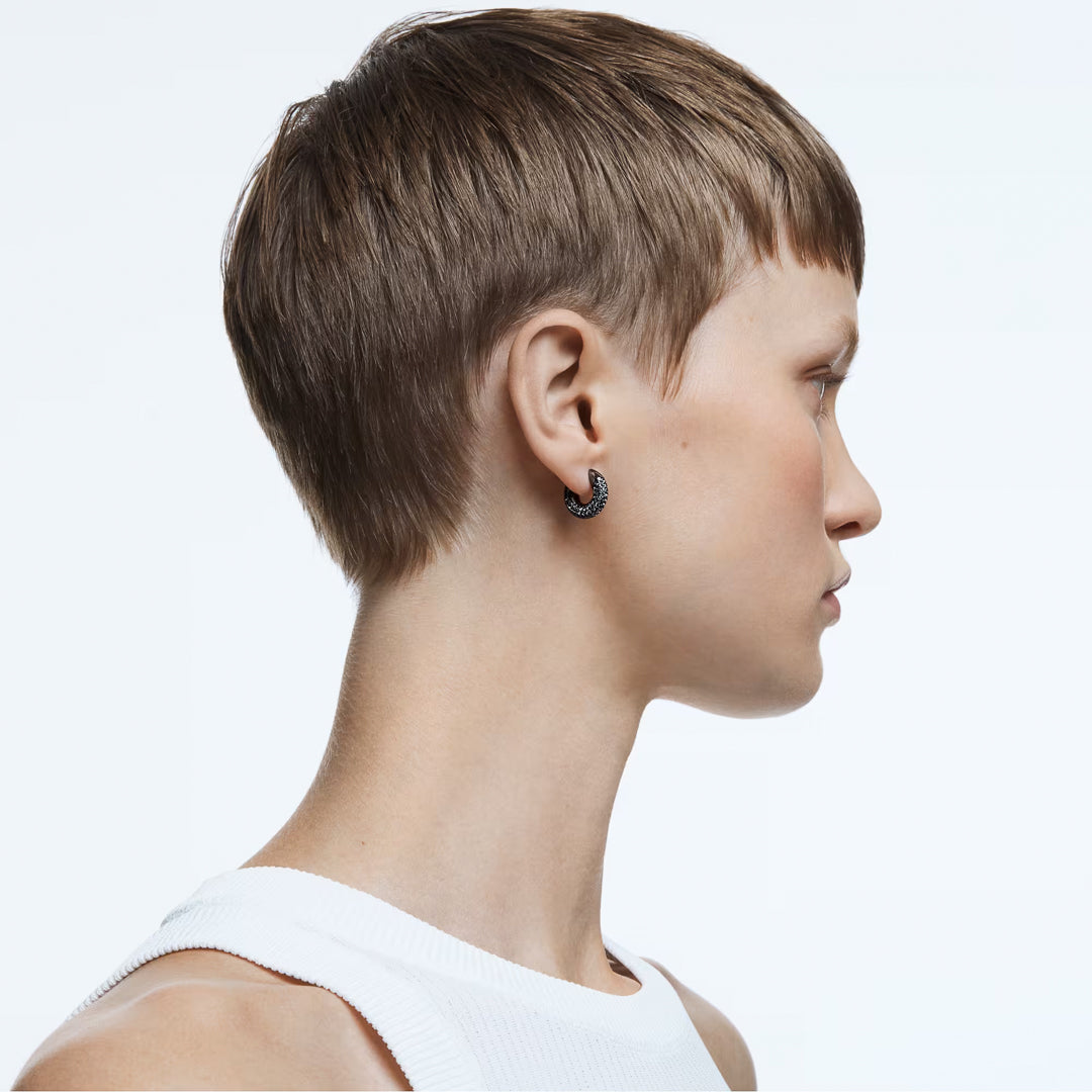 A person with short brown hair is shown in profile, wearing a white sleeveless top. They are adorned with Swarovski Dextera Hoop Earrings and are standing against a plain, light background.
