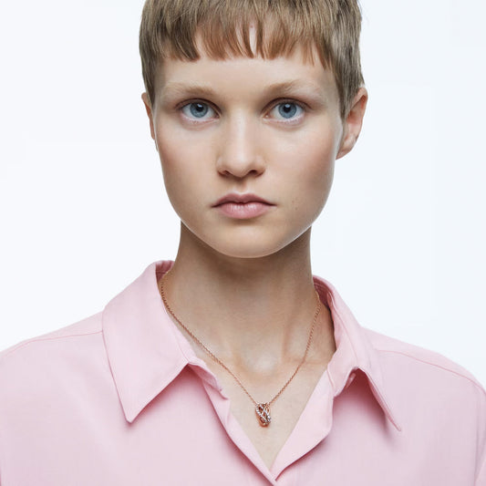 A person with short hair wears a light pink collared shirt and an elegant Swarovski Hyperbola Necklace Pendant in a rose-gold tone, featuring a zirconia heart pendant. The background is plain white, and the person's expression is neutral.