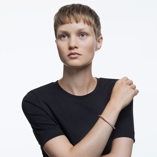 A person with short light brown hair wearing a black t-shirt poses against a light background. Their left arm is raised slightly, showcasing the delicate Swarovski Hyperbola Bracelet in rose-gold on their wrist. They are looking slightly upward.