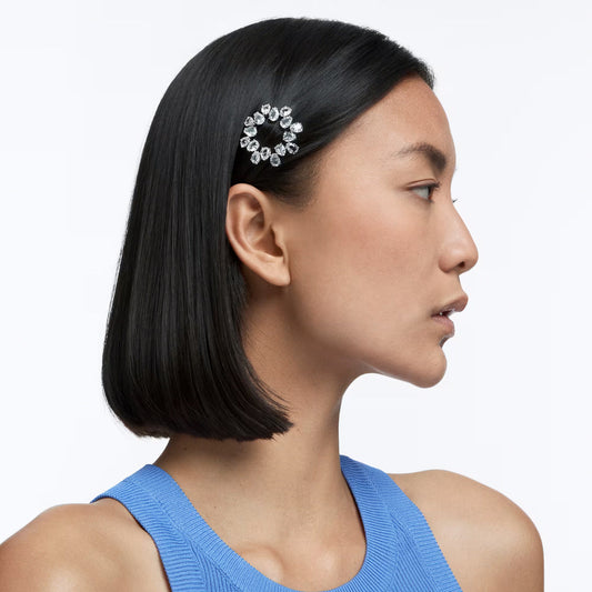A person with sleek black hair is shown in profile against a simple white backdrop, elegantly accessorized with the Swarovski Millenia Hair Clip, beautifully embellished with pear-cut crystals. They are attired in a stylish blue sleeveless top.