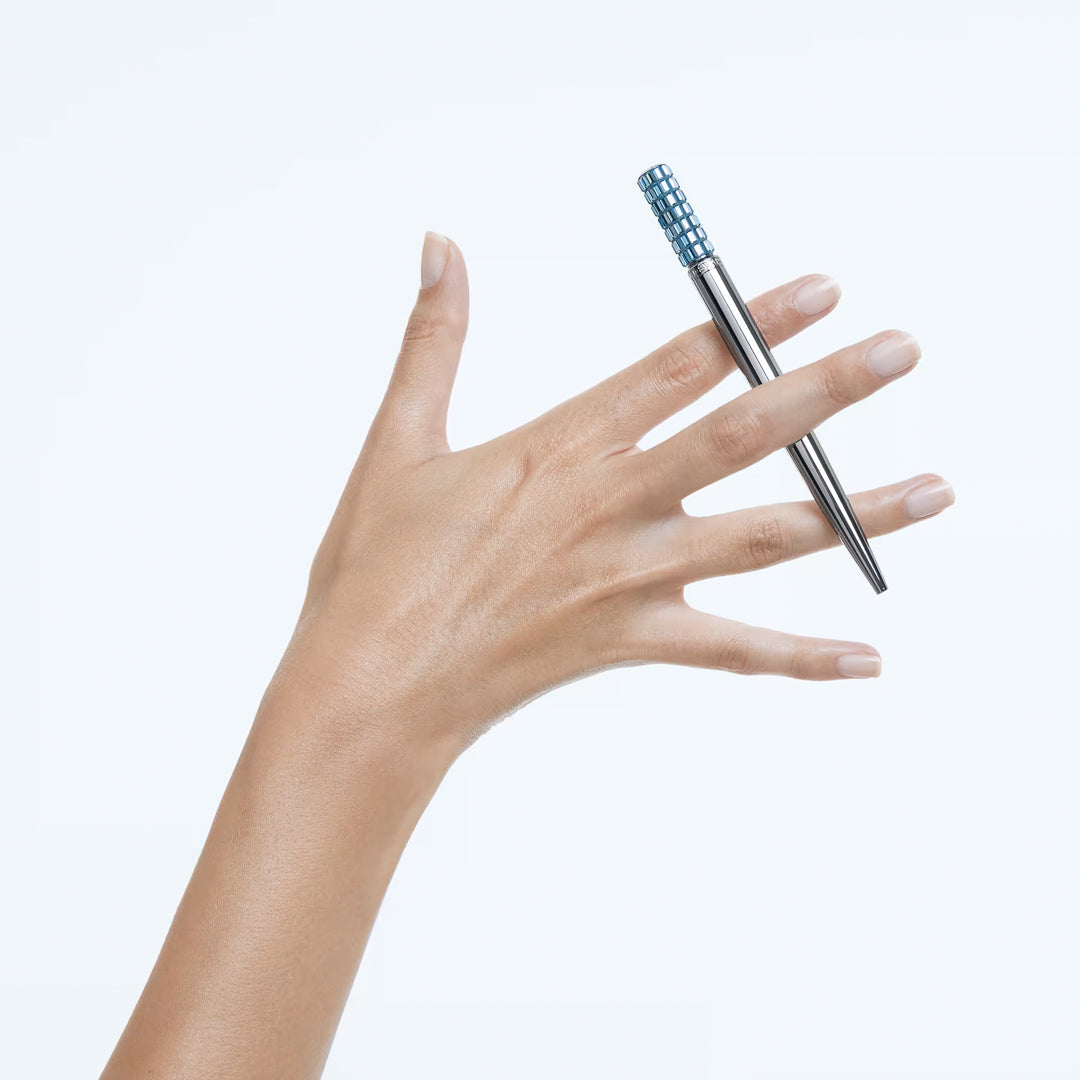 A hand elegantly balancing a Swarovski Ballpoint Pen adorned with shimmering crystals against a plain white background, featuring a distinct textured blue grip near the top.