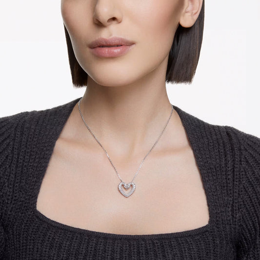 A person with short hair wears a black textured sweater, highlighting the Swarovski Sublima Heart Necklace Pendant, which features a double heart design adorned with pavé crystals. The necklace stands out beautifully against the plain white background.