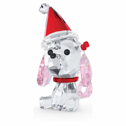 Swarovski's Holiday Cheers Poodle Home Décor is a charming crystal figurine depicting a poodle with pink ears, a red collar, and a festive Santa hat. This delightful piece is adorned with a red bow on its head and showcases classic black eyes and nose, captured in an endearing playful pose.