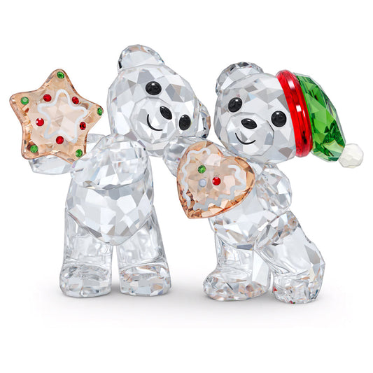 A pair of dazzling crystal teddy bears from Swarovski glimmer with their sparkling facets. One bear, part of the Kris Bear collection and known as the Christmas 2022 Annual Edition Home Décor piece, holds a star-shaped ornament, while its companion clutches a heart-shaped one. Both bears are decorated with red and green accents, with the bear on the right wearing festive hats, creating an enchanting and delightful holiday display.