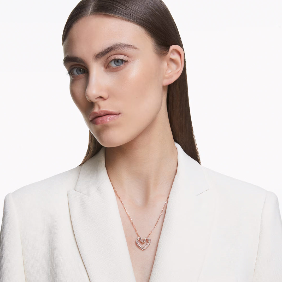 A person with long brown hair is wearing a white blazer and the Swarovski Sublima Necklace Pendant, which features a heart-shaped design adorned with pavé crystals. The plain white background accentuates the elegant and minimalist style, highlighted by the subtle sparkle of Swarovski's craftsmanship.