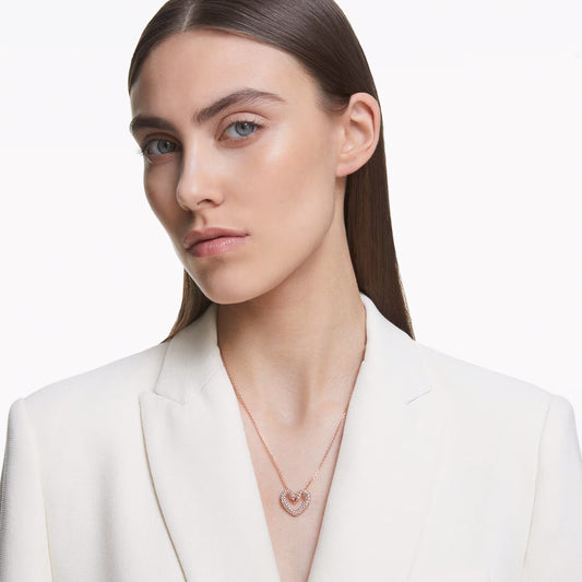 A person with long brown hair is wearing a white blazer and the Swarovski Sublima Necklace Pendant, which features a heart-shaped design adorned with pavé crystals. The plain white background accentuates the elegant and minimalist style, highlighted by the subtle sparkle of Swarovski's craftsmanship.