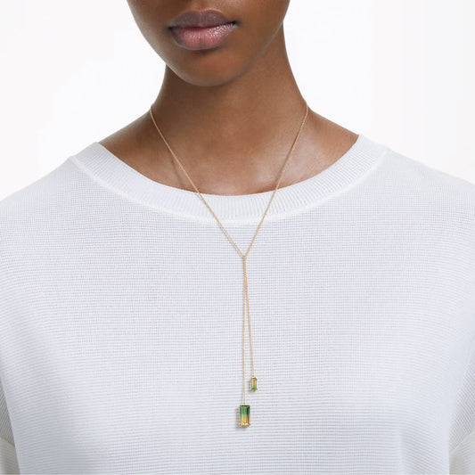 A person is adorned in a white textured top and the Swarovski Millenia Y Necklace Pendant by Swarovski, featuring two gold-tone plated pendants with green gemstones of varying sizes and lengths, creating a minimalist and elegant appearance.