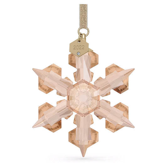 A Swarovski Festive2022 Annual Edition Ornament Home Décor piece elegantly adorns a space with its light amber-hued crystal snowflake design, hanging from a gold ribbon. It showcases intricate facets surrounding a hexagonal center and is adorned with a small gold tag engraved "2022" and a gemstone accent, making it perfect for holiday decorations.